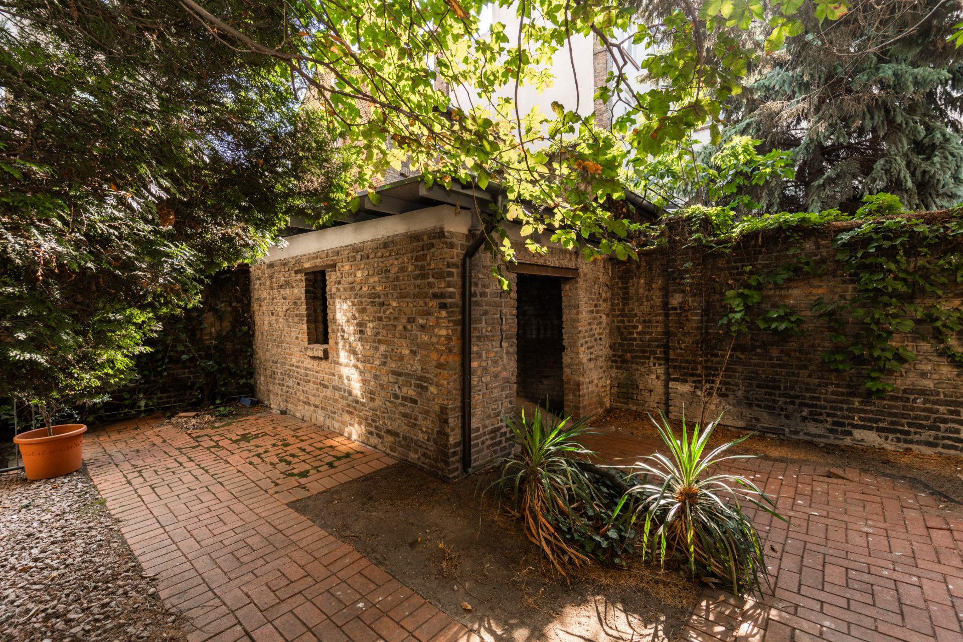 Kleines Backsteingebäude im Innenhof mit Pflastersteinen, Pflanzen und Kletterpflanzen an der Mauer.