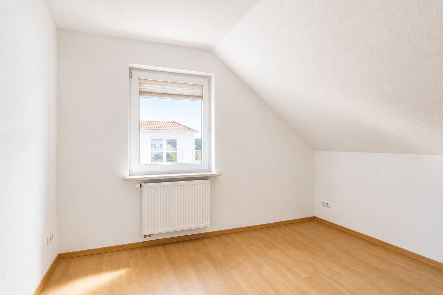 Leeres Zimmer mit Dachschräge, Fenster, Heizkörper und hellem Holzboden.