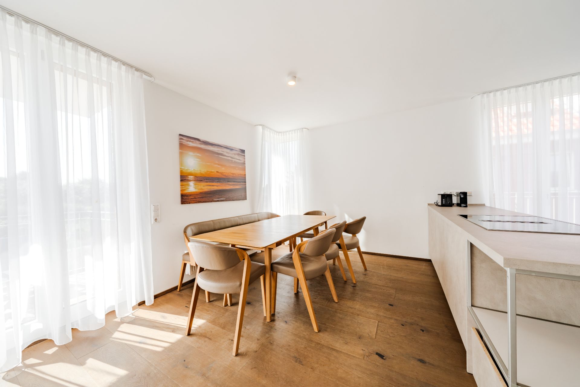 Heller Essbereich mit Holztisch, sechs Stühlen, Küchenzeile und großem Fenster mit weißen Vorhängen.