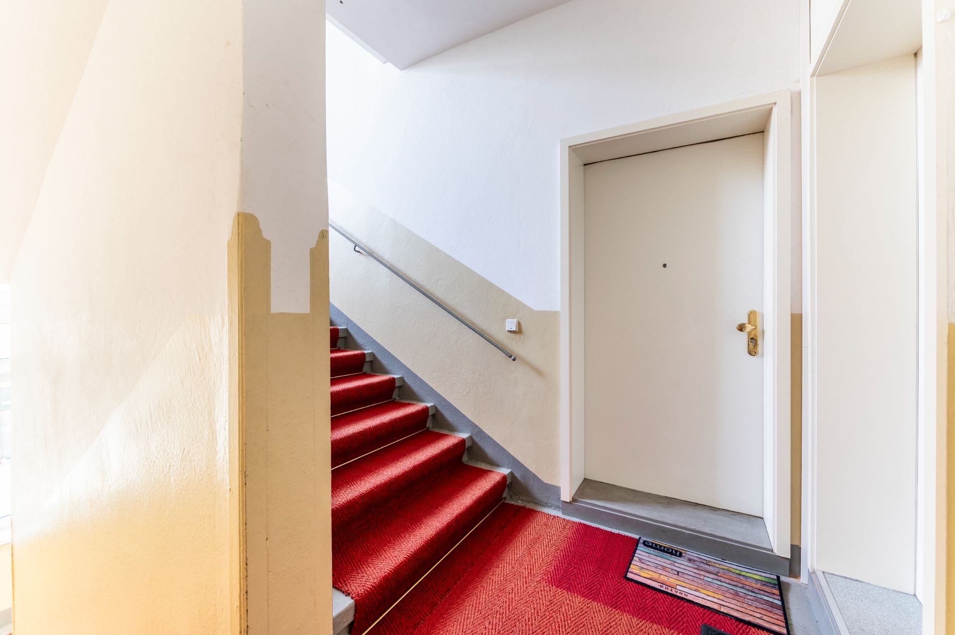 Treppenhaus mit rotem Teppich und weißer Wohnungstür, kleiner bunter Teppich vor der Tür.