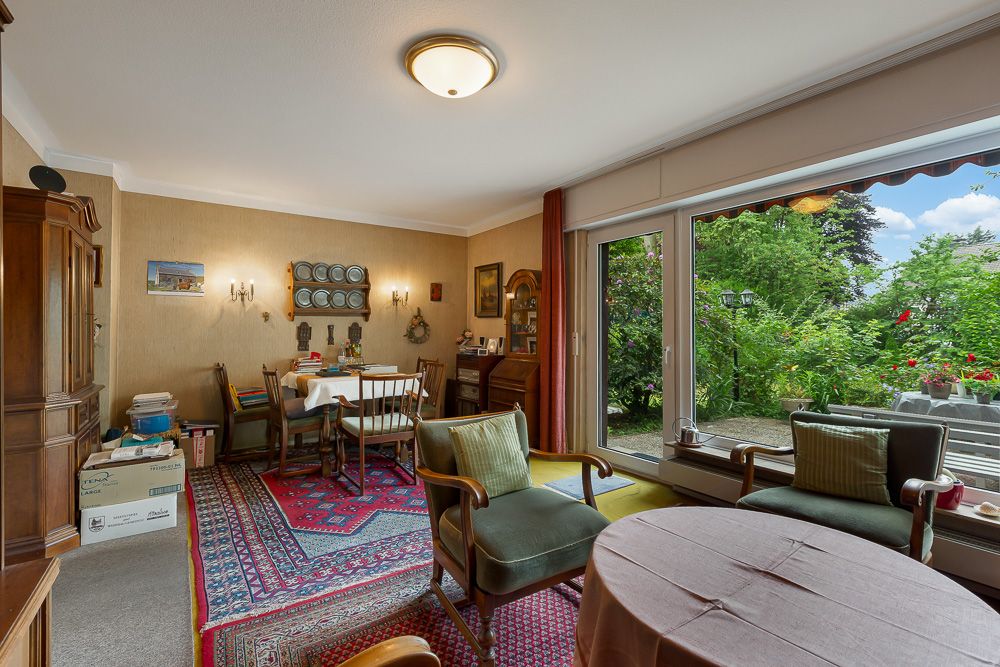 Wohnzimmer mit Teppich, mehreren Sitzgelegenheiten, großem Fenster und Blick in einen grünen Garten.