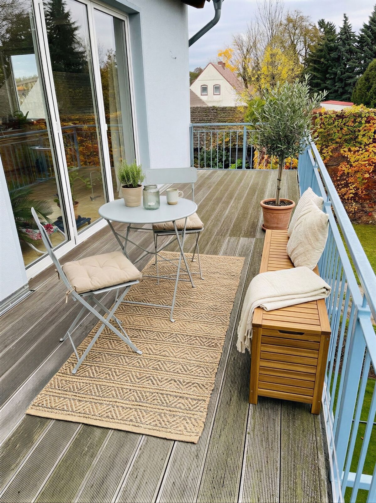 Balkon mit Holzboden, kleinem Tisch, zwei Stühlen, Bank, Pflanze und Teppich.