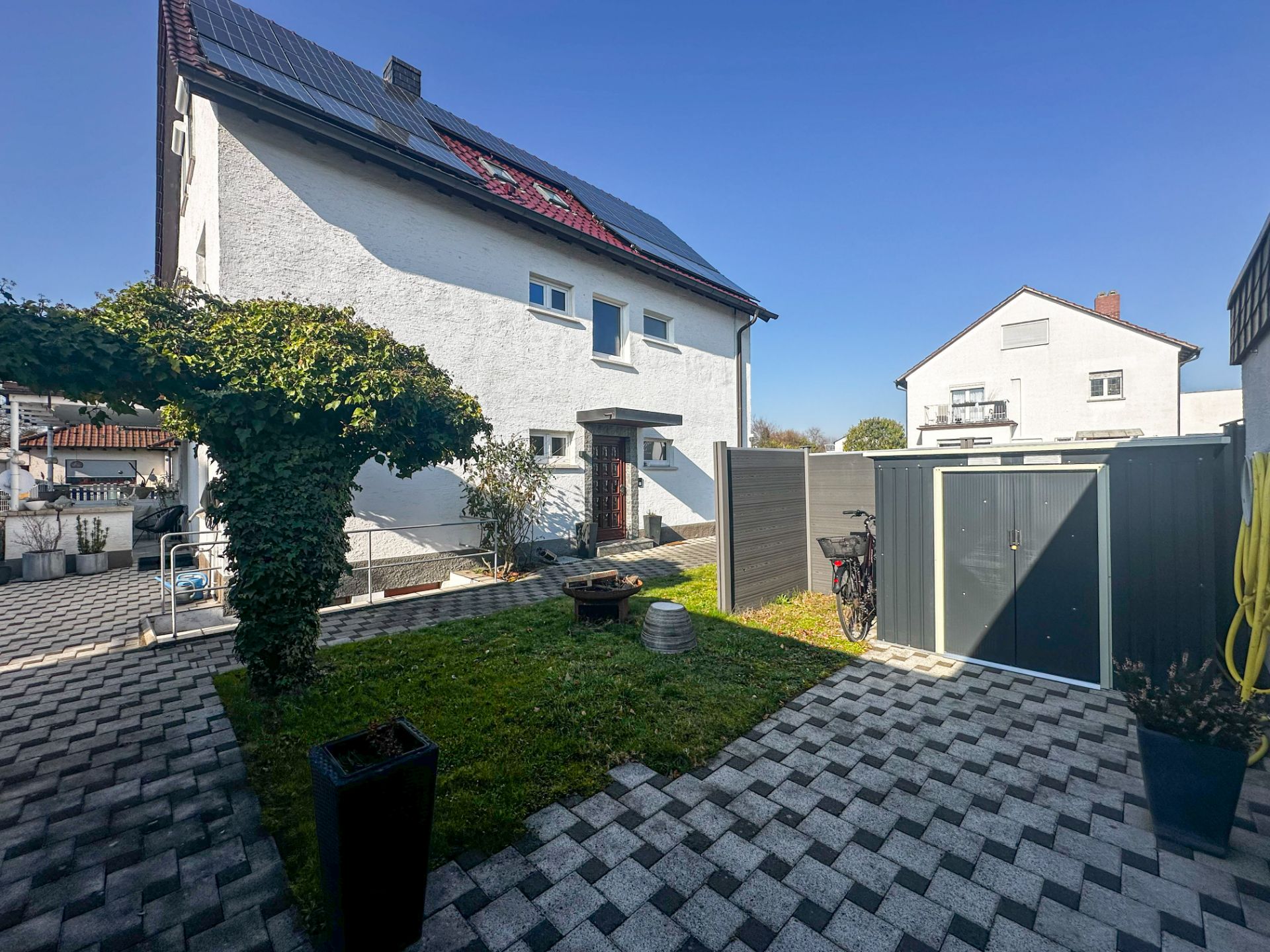 Garten mit gepflastertem Weg, Baum, Schuppen und weißem Wohnhaus im Hintergrund.