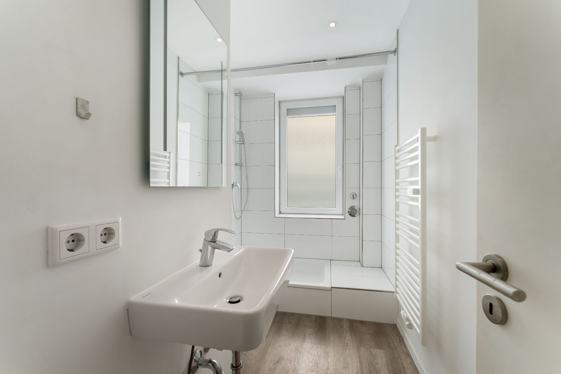 Helles Badezimmer mit Fenster, Waschbecken, Dusche und Handtuchheizkörper.