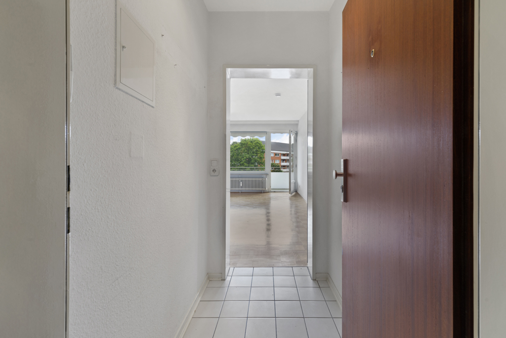 Blick vom Flur mit Fliesenboden in ein helles Wohnzimmer mit großen Fenstern.
