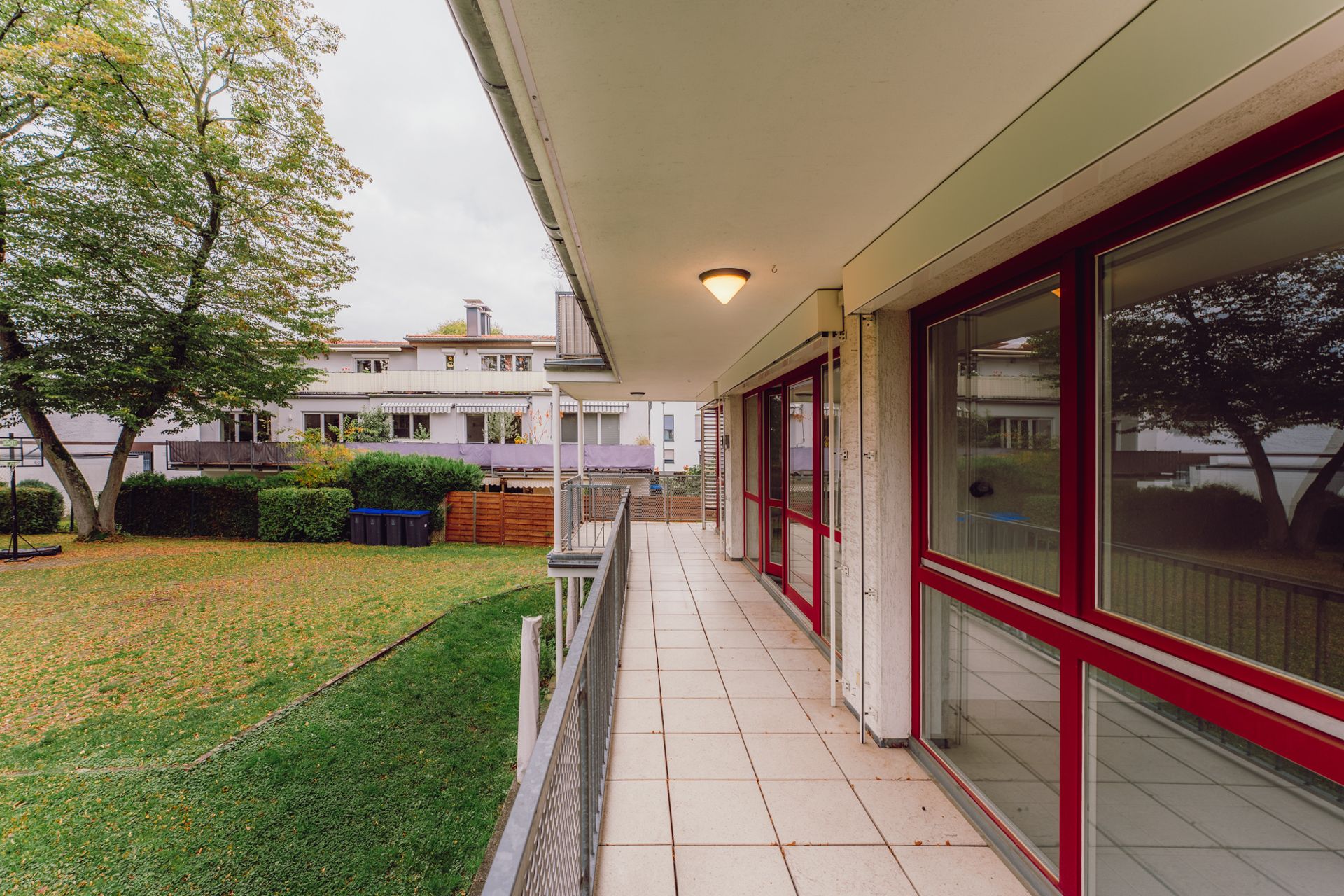 Überdachter Balkon mit rot gerahmten Fenstern, Blick auf Garten und Wohnhäuser.