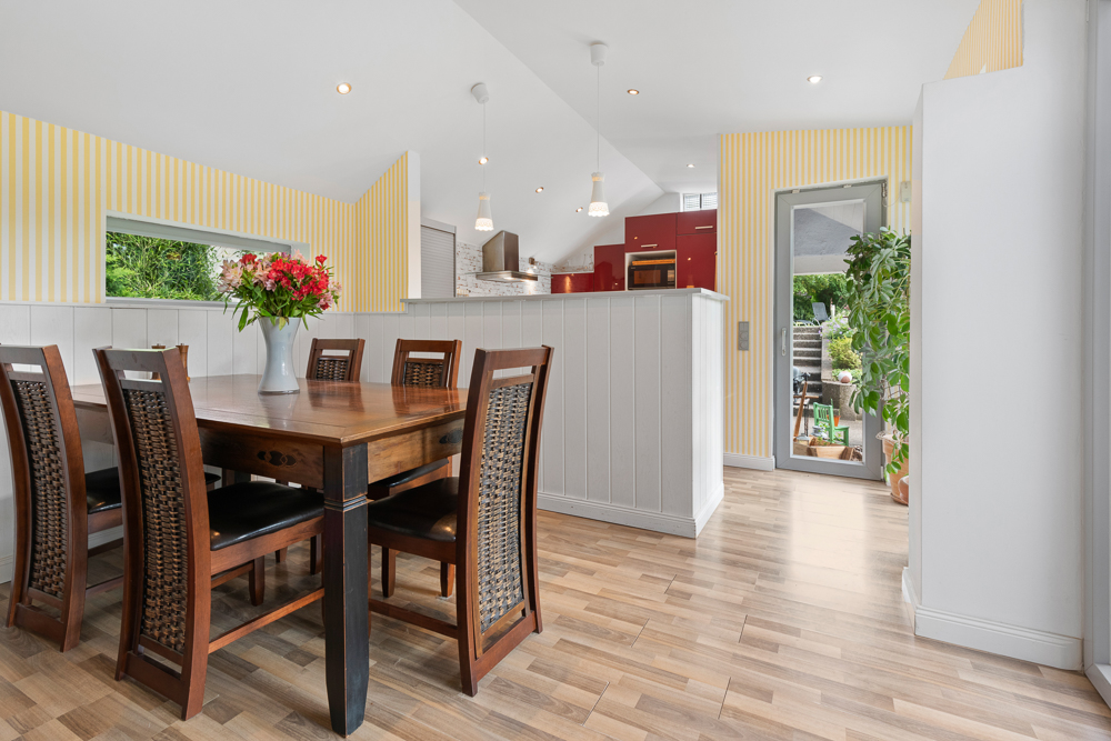 Esszimmer mit Holztisch, sechs Stühlen, Blumenstrauß und Blick in offene Küche, helle Wände, Holzfußboden.