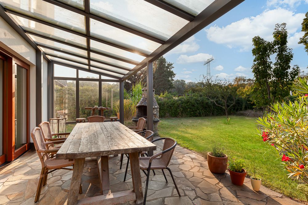 Überdachte Terrasse mit Holztisch und Stühlen, Blick auf Garten mit Rasen und Pflanzen.