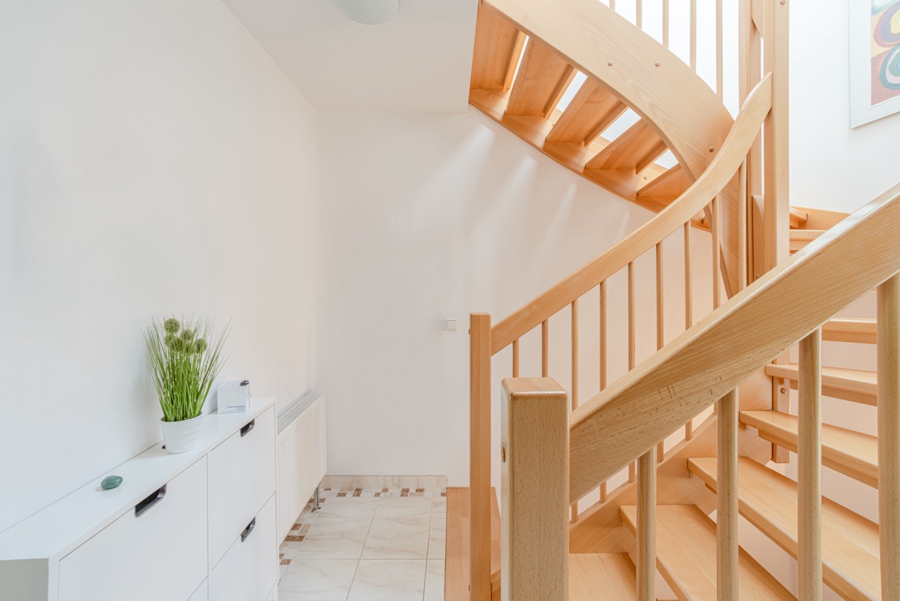 Heller Flur mit Holztreppe, weißem Schuhschrank und Topfpflanze auf Fliesenboden.