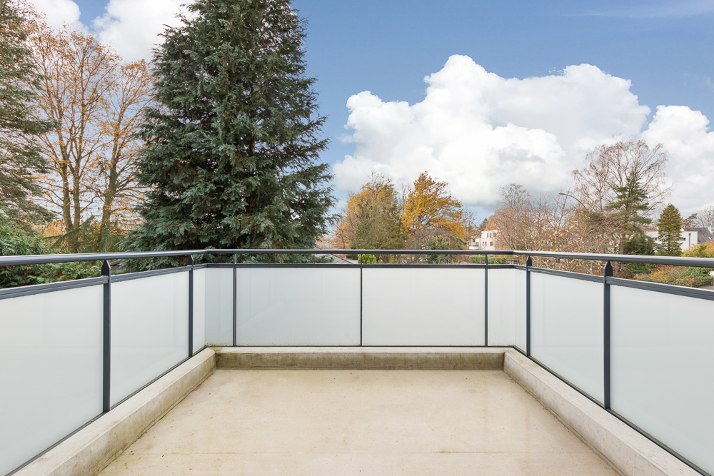 Leerer Balkon mit Glasgeländer und Blick auf Bäume und Häuser im Hintergrund.