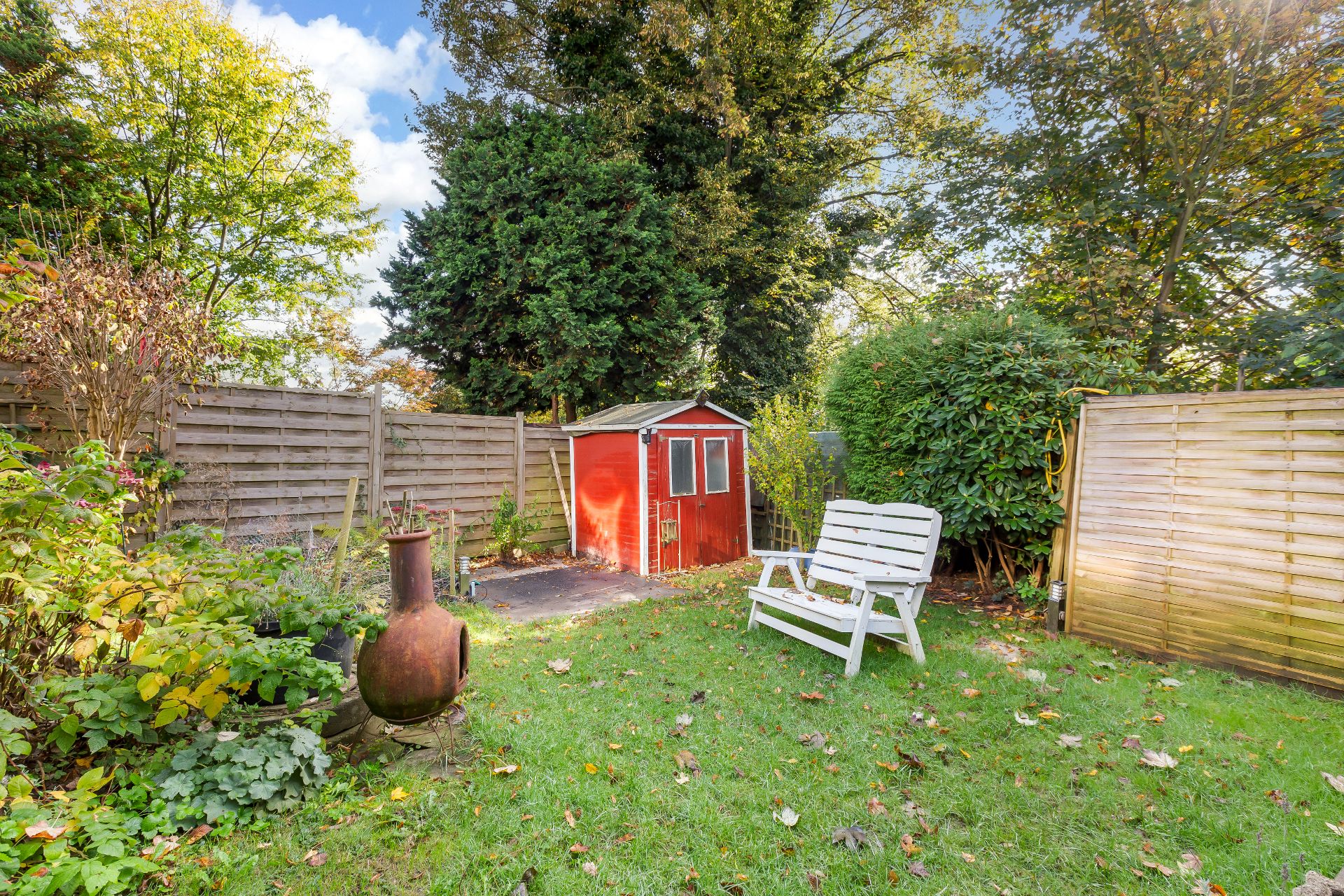 Garten mit rotem Geräteschuppen, weißer Bank und Kaminofen, umgeben von Holzzaun und Büschen.