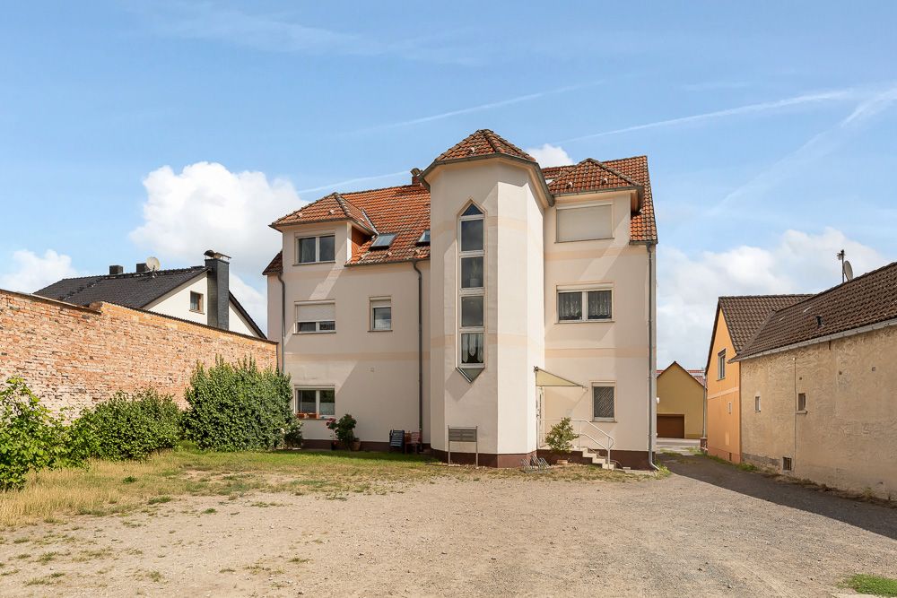 Mehrstöckiges Wohnhaus mit rotem Dach und mehreren Fenstern, davor ein Hof mit Kiesfläche.