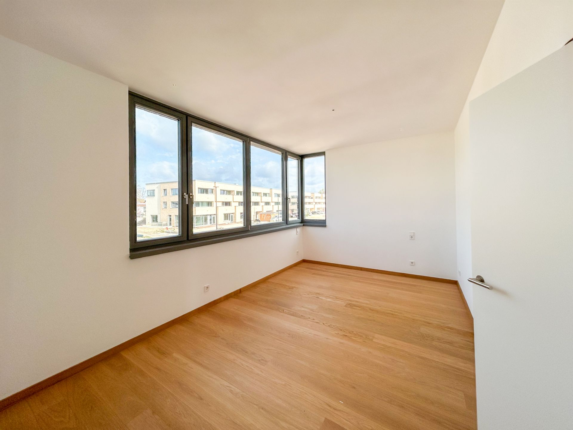 Helles, leeres Zimmer mit großem Eckfenster und Parkettboden.