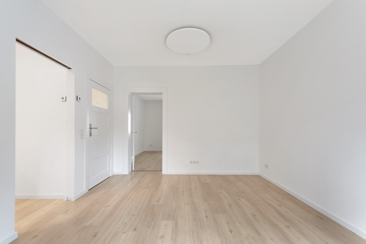 Heller, leerer Raum mit Holzboden, weißen Wänden und Türen, Deckenlampe in der Mitte.