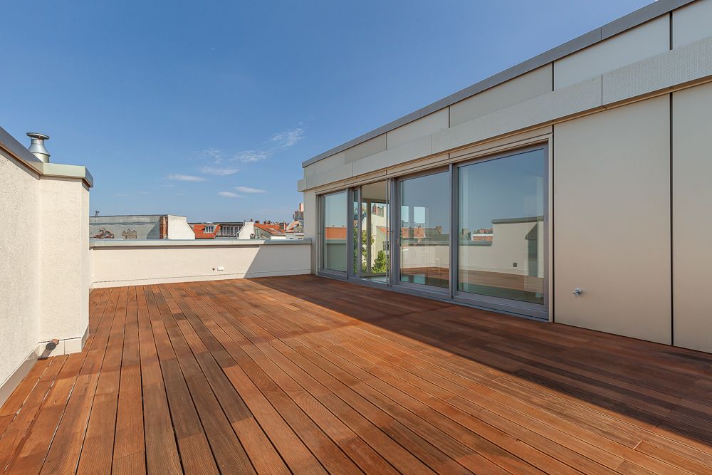 Große Dachterrasse mit Holzboden und bodentiefen Fenstern, Blick auf Dächer unter blauem Himmel.