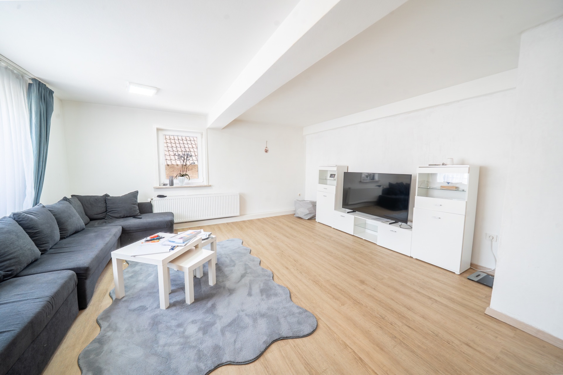 Helles Wohnzimmer mit grauem Sofa, Teppich, Couchtisch und großem Fernseher auf Holzfußboden.