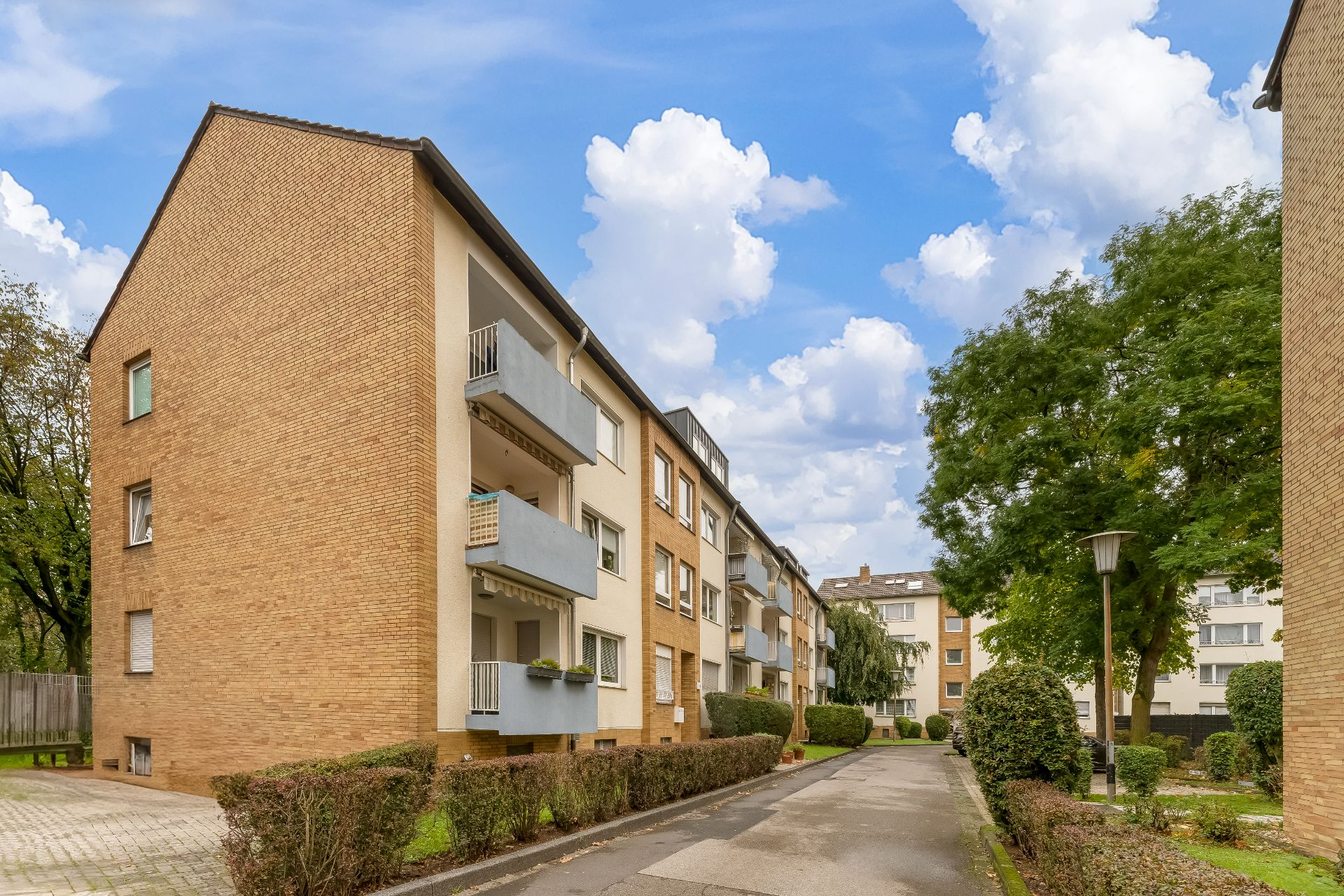 Mehrstöckiges Wohnhaus mit Balkonen, umgeben von Büschen und Bäumen unter blauem Himmel.