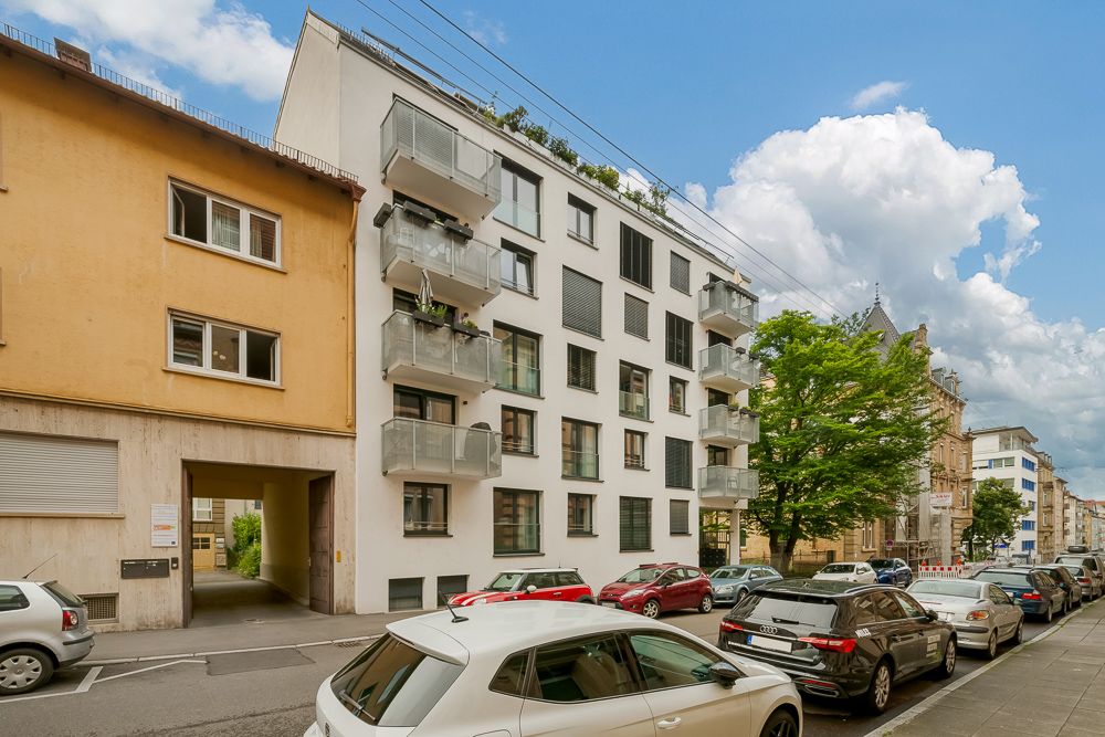 Mehrfamilienhaus mit Balkonen an einer Straße mit parkenden Autos und Bäumen.