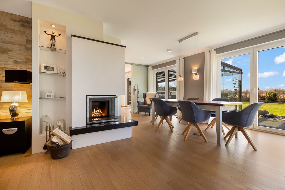 Modernes Wohnzimmer mit Kamin, Esstisch, blauen Stühlen und großen Fenstern mit Blick in den Garten.