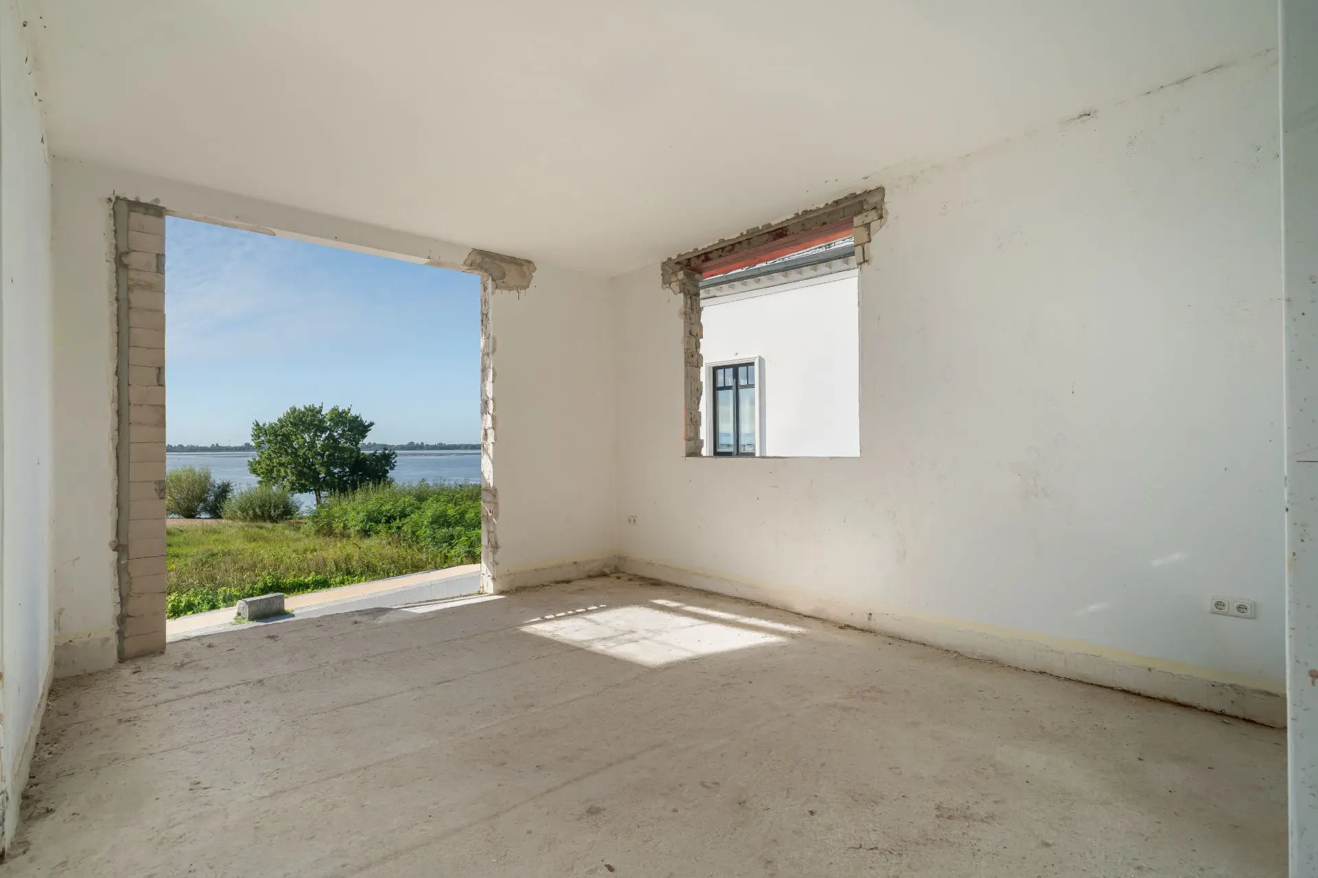 Leerer Raum mit unverputzten Fensteröffnungen und Blick ins Grüne und auf Wasser.
