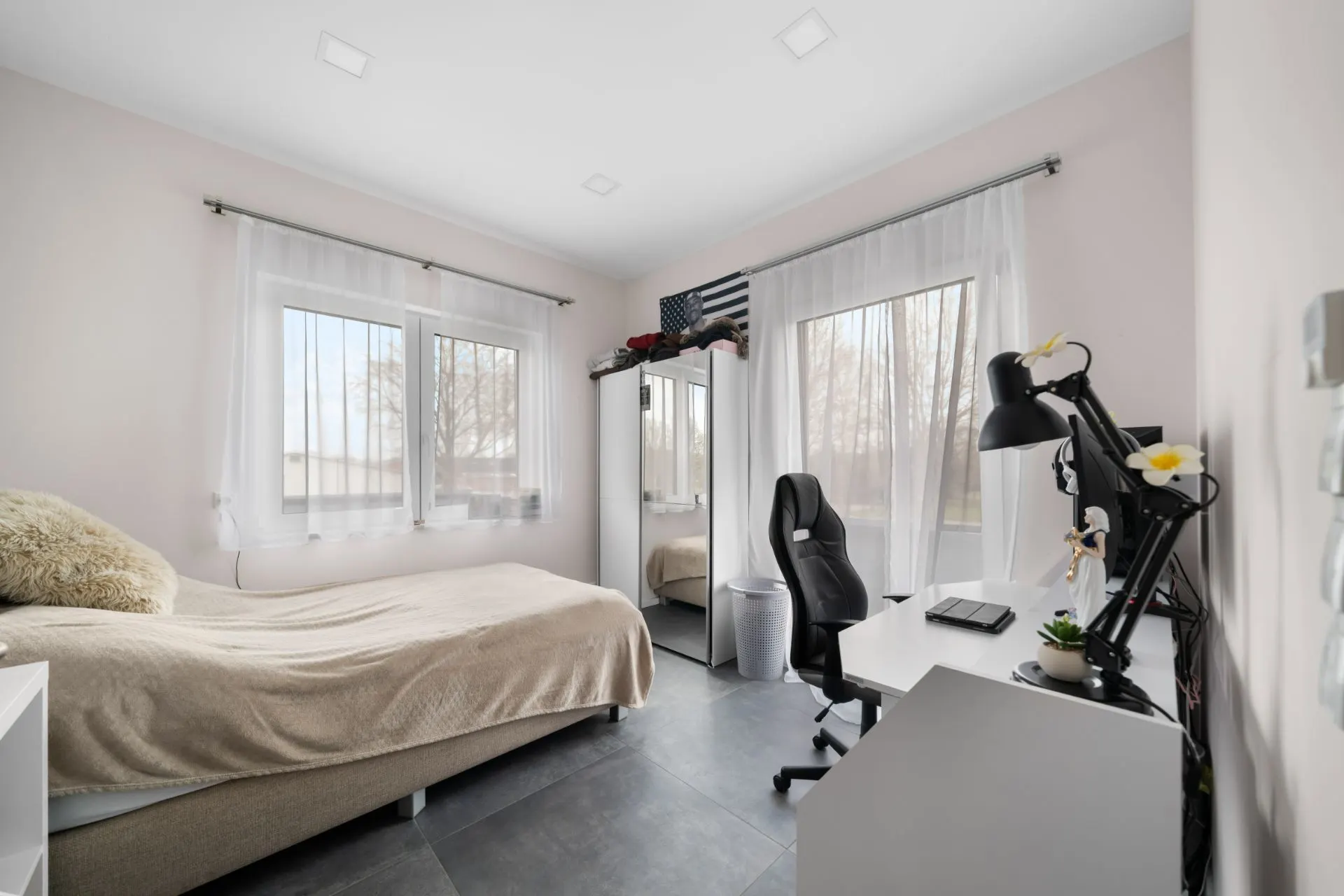Helles Schlafzimmer mit Bett, Schreibtisch, Stuhl und Spiegel-Schrank neben zwei Fenstern.