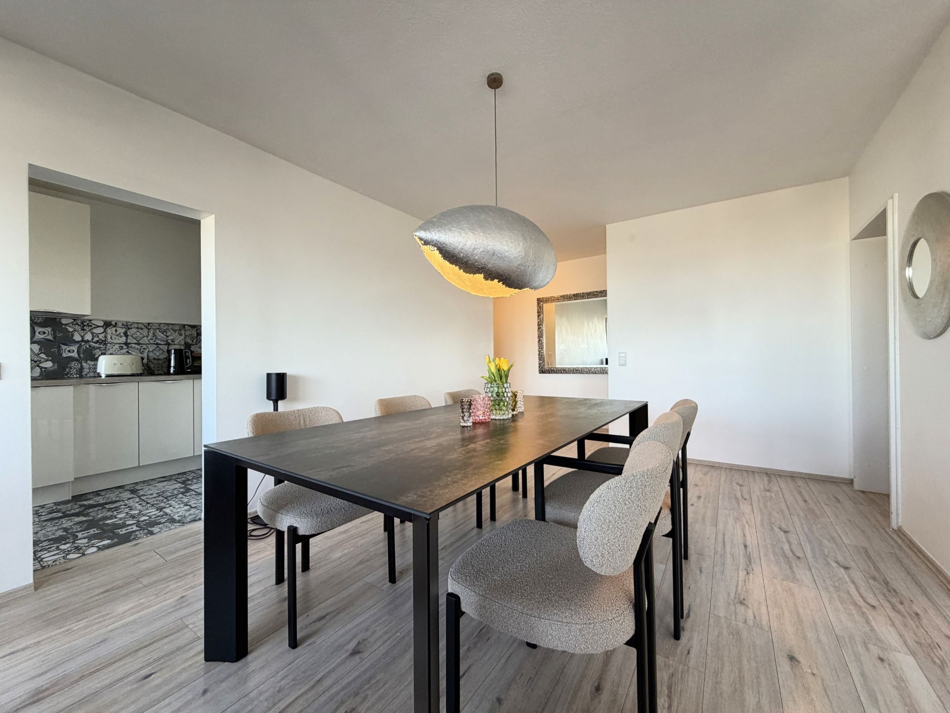 Esszimmer mit schwarzem Tisch, sechs Stühlen und großer Hängelampe. Zugang zur Küche sichtbar.