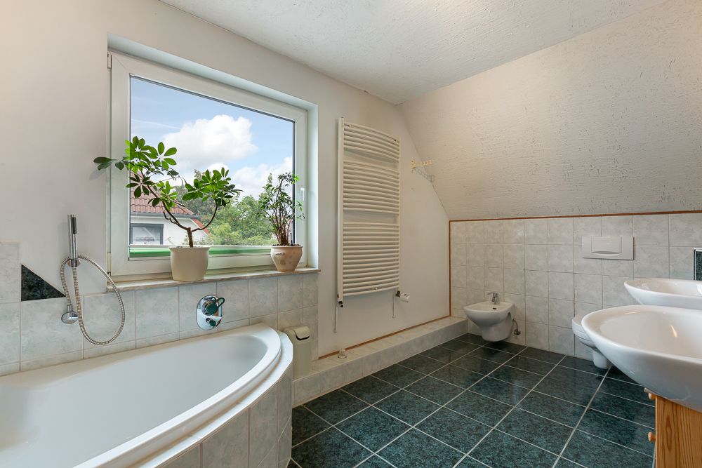 Helles Badezimmer mit Badewanne, zwei Waschbecken, Bidet, Pflanzen am Fenster und grünem Fliesenboden.