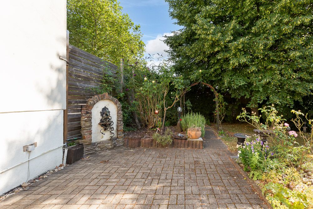 Gepflasterte Terrasse mit Pflanzen, kleinem Brunnen und angrenzendem Gartenbereich.
