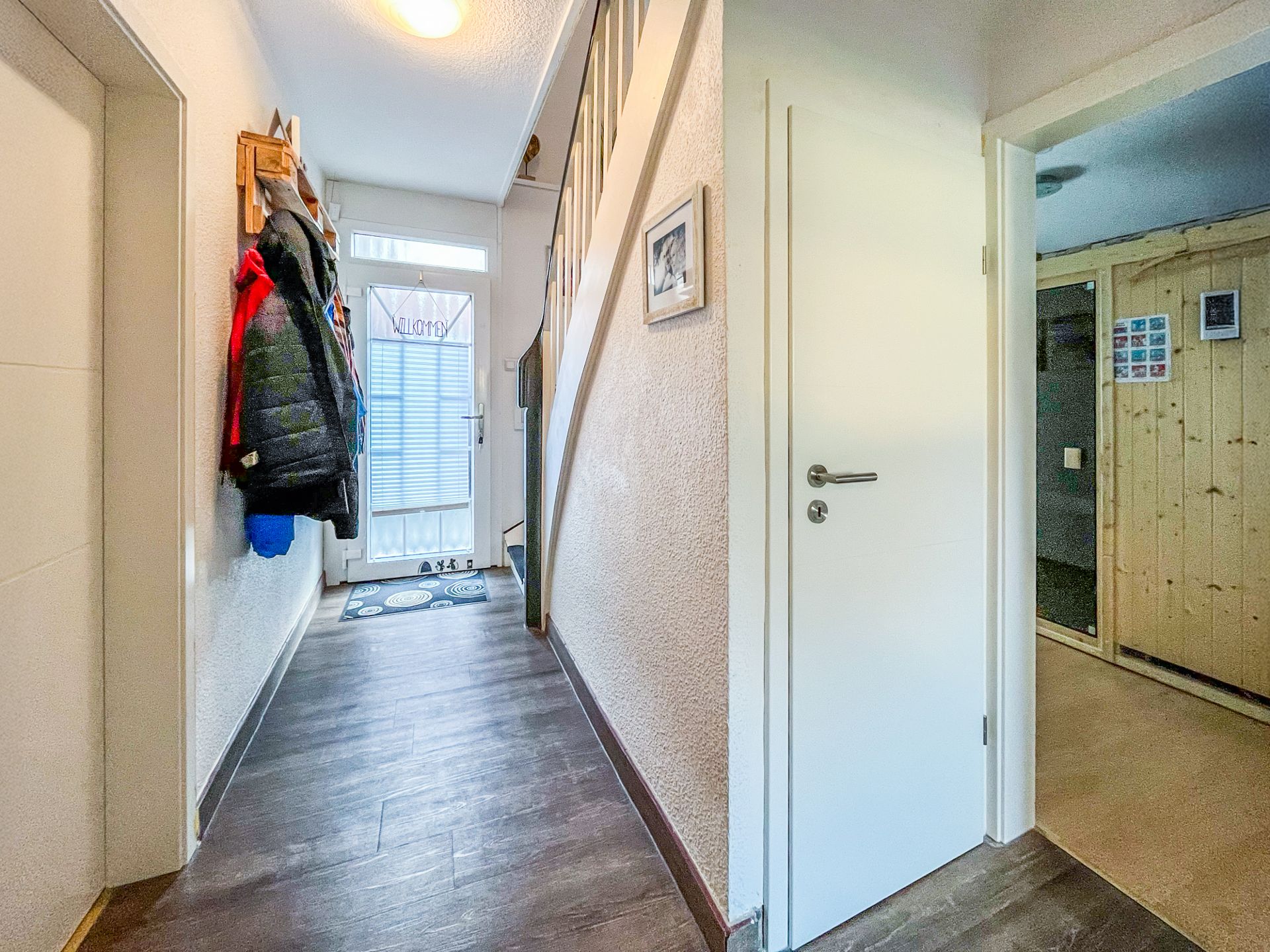 Heller Flur mit Garderobe, Treppe, Tür und Blick auf einen Raum mit Holzverkleidung.