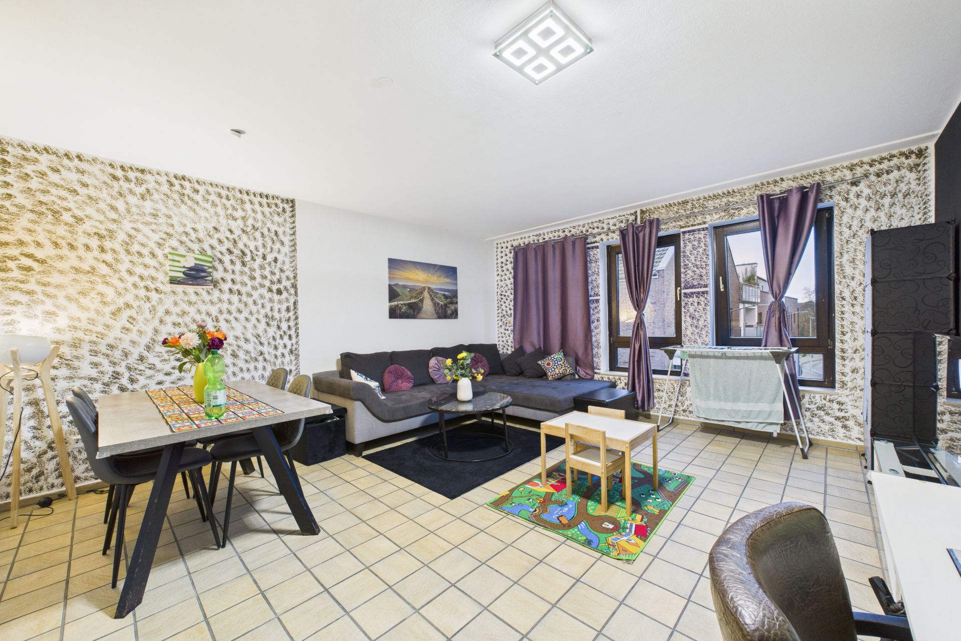 Helles Wohnzimmer mit Ecksofa, Esstisch, Spielteppich und großen Fenstern mit lila Vorhängen.