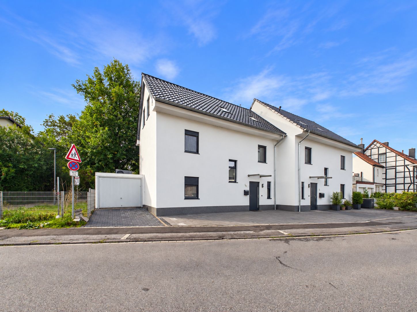 Modernes, weißes Doppelhaus mit schwarzem Dach, Garage und Gehweg an einer Straße.