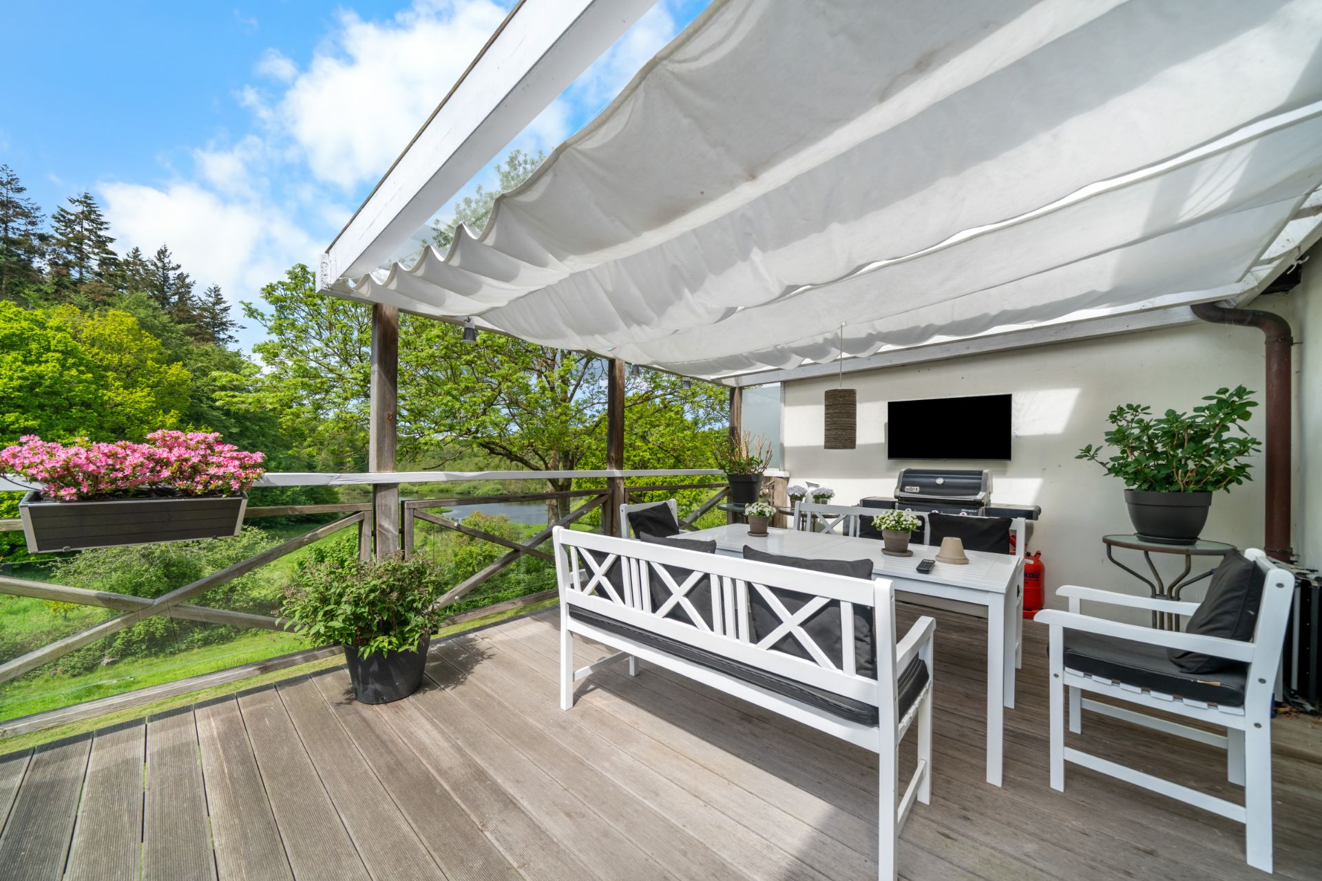 Überdachte Holzterrasse mit weißen Gartenmöbeln, Grill und Blumentöpfen, umgeben von viel Grün.