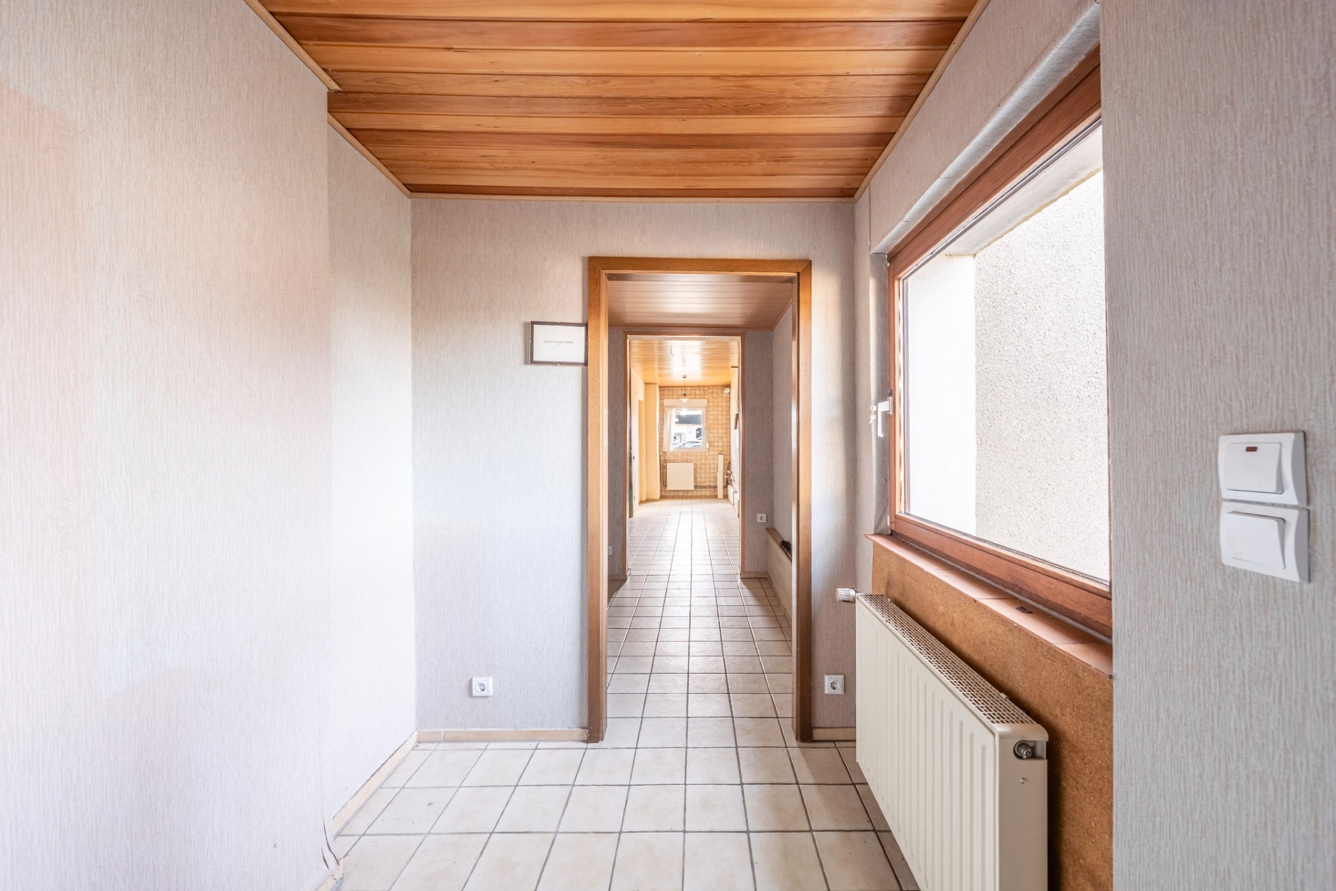 Heller Flur mit Fliesenboden, Holzdecke und Fenster auf der rechten Seite.