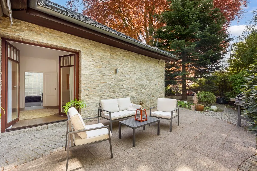 Terrasse mit Sitzgruppe und Tisch neben einem Haus mit Steinmauer, umgeben von Bäumen und Pflanzen.