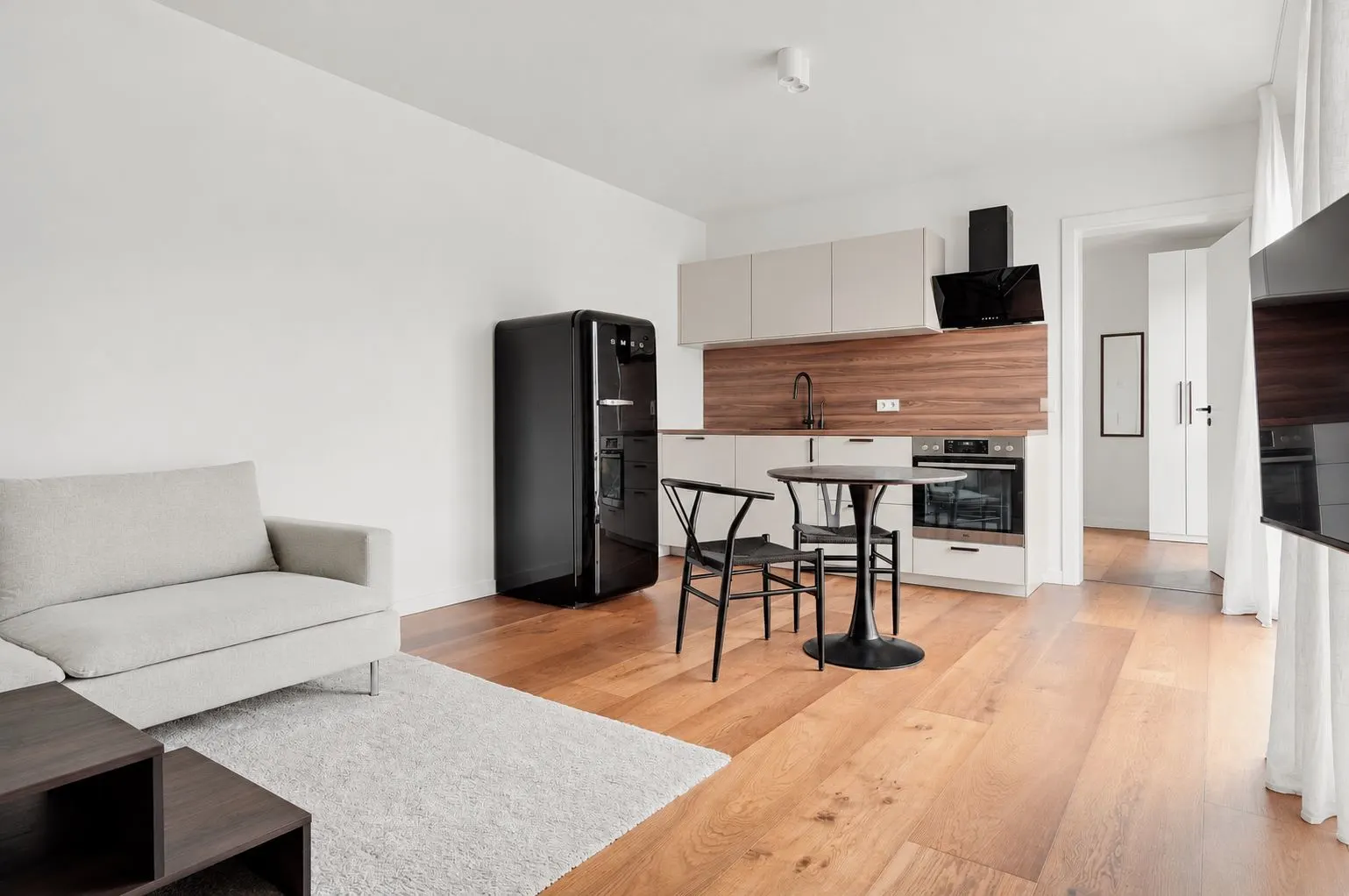 Helles Wohnzimmer mit Sofa, Teppich, Esstisch, offener Küche und schwarzem Kühlschrank.
