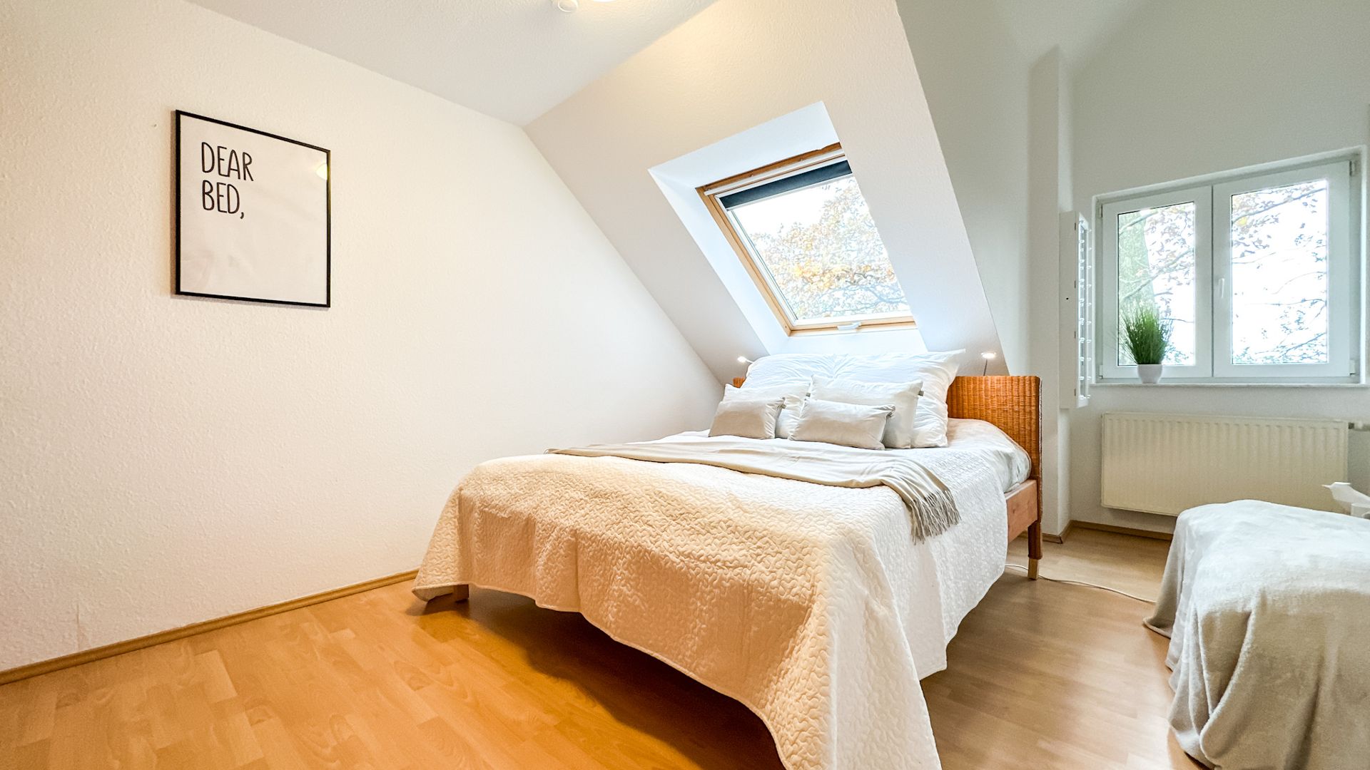 Helles Schlafzimmer mit Doppelbett, Dachfenster, Sessel und Fenster mit Pflanze.