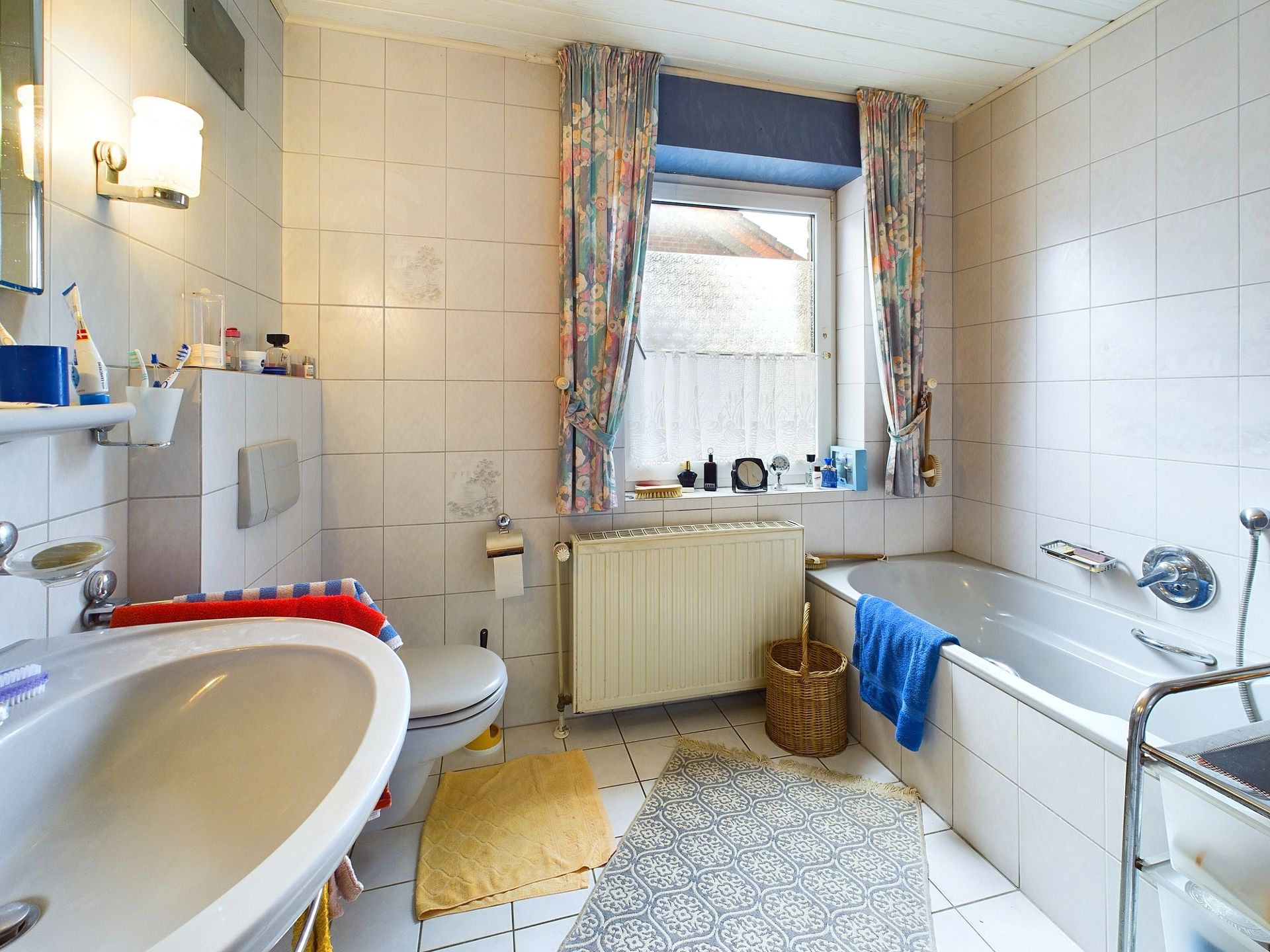 Helles Badezimmer mit Badewanne, Waschbecken, Toilette und Fenster mit bunten Vorhängen.