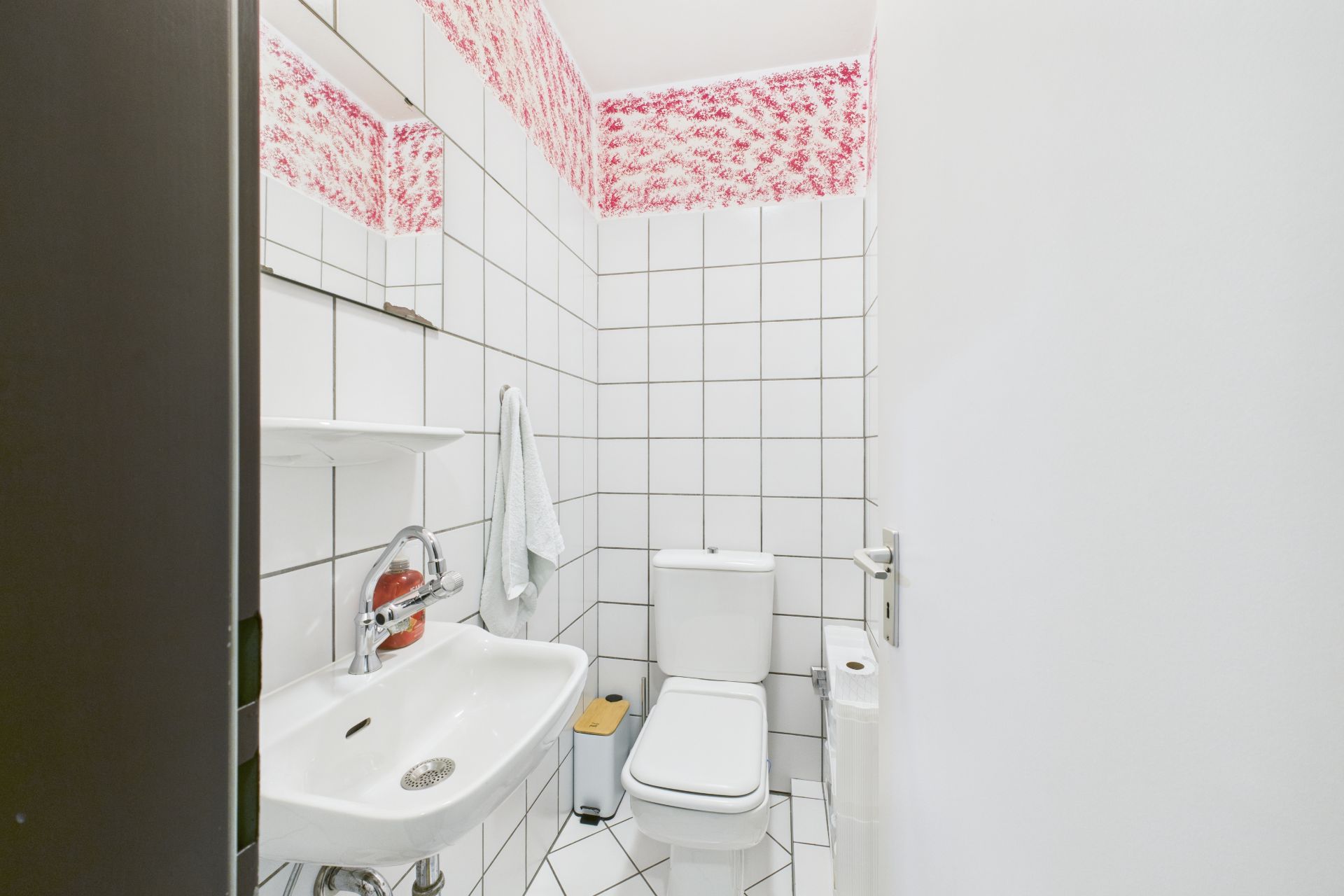 Kleines Bad mit weiß gefliesten Wänden, WC, Waschbecken und pink gemustertem Streifen an der Decke.