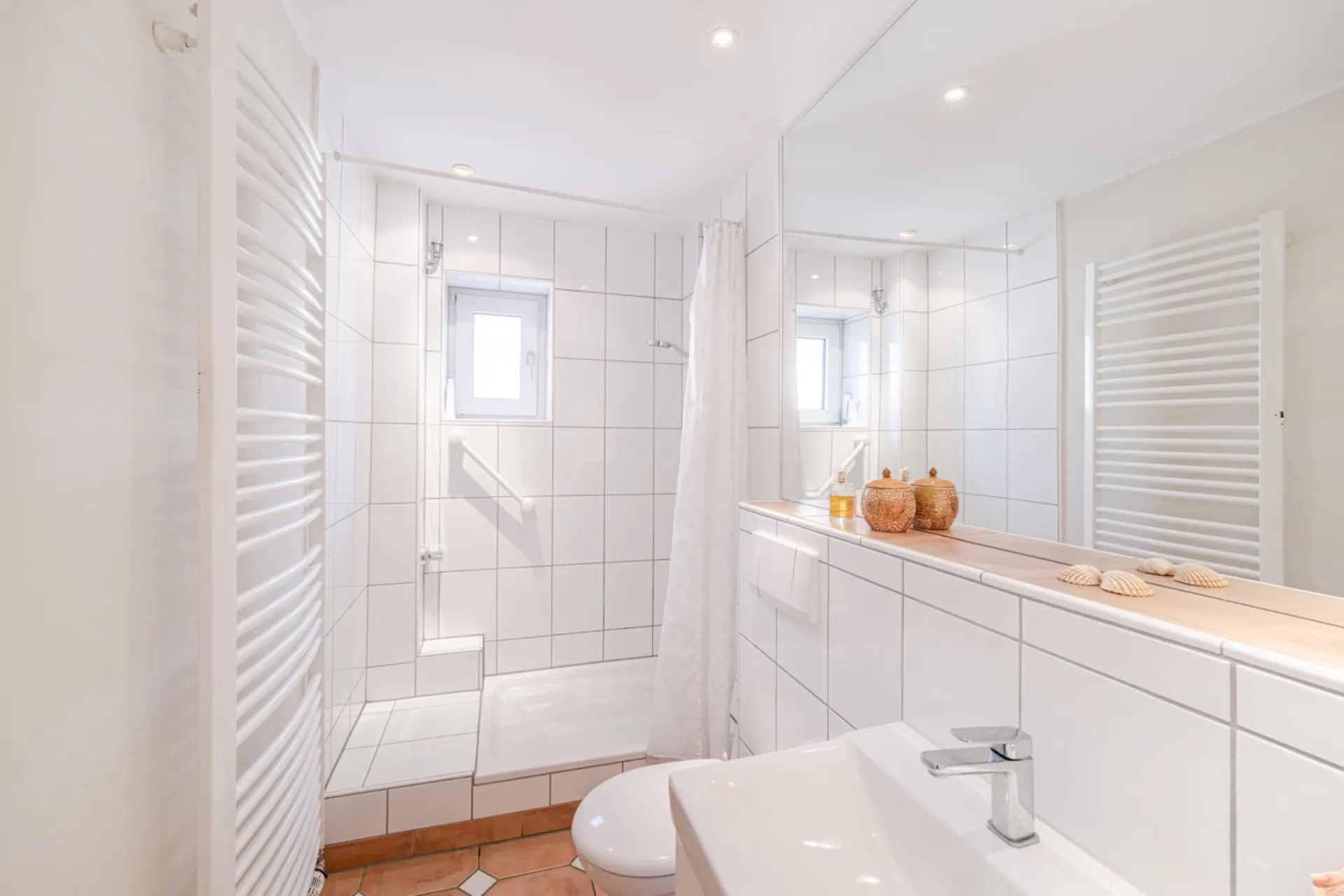 Helles Badezimmer mit Dusche, Waschbecken, WC und dekorativen Muscheln auf dem Regal.