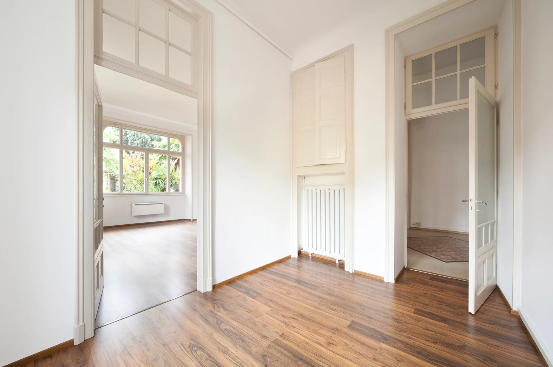 Heller Raum mit Holzboden, weißen Wänden und großen Fenstern, Türen zu weiteren Zimmern.