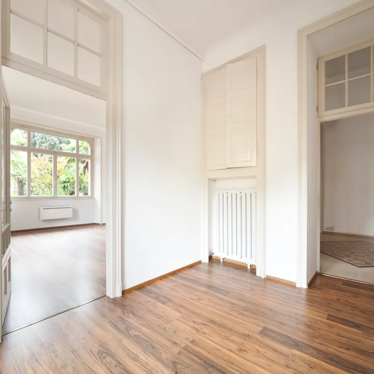 Heller Raum mit Holzboden, weißen Wänden und großen Fenstern, Türen zu weiteren Zimmern.