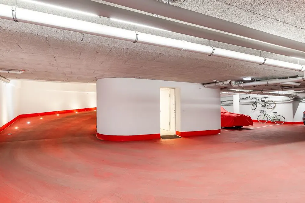 Helle Tiefgarage mit rotem Boden, Tür in der Wand, abgedecktem Auto und aufgehängten Fahrrädern.