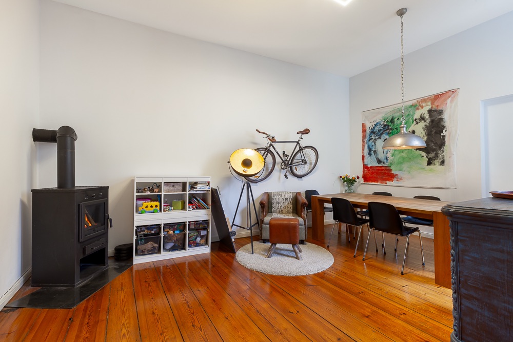 Heller Wohnraum mit Holzofen, Bücherregal, Sessel, Esstisch und Fahrrad an der Wand.