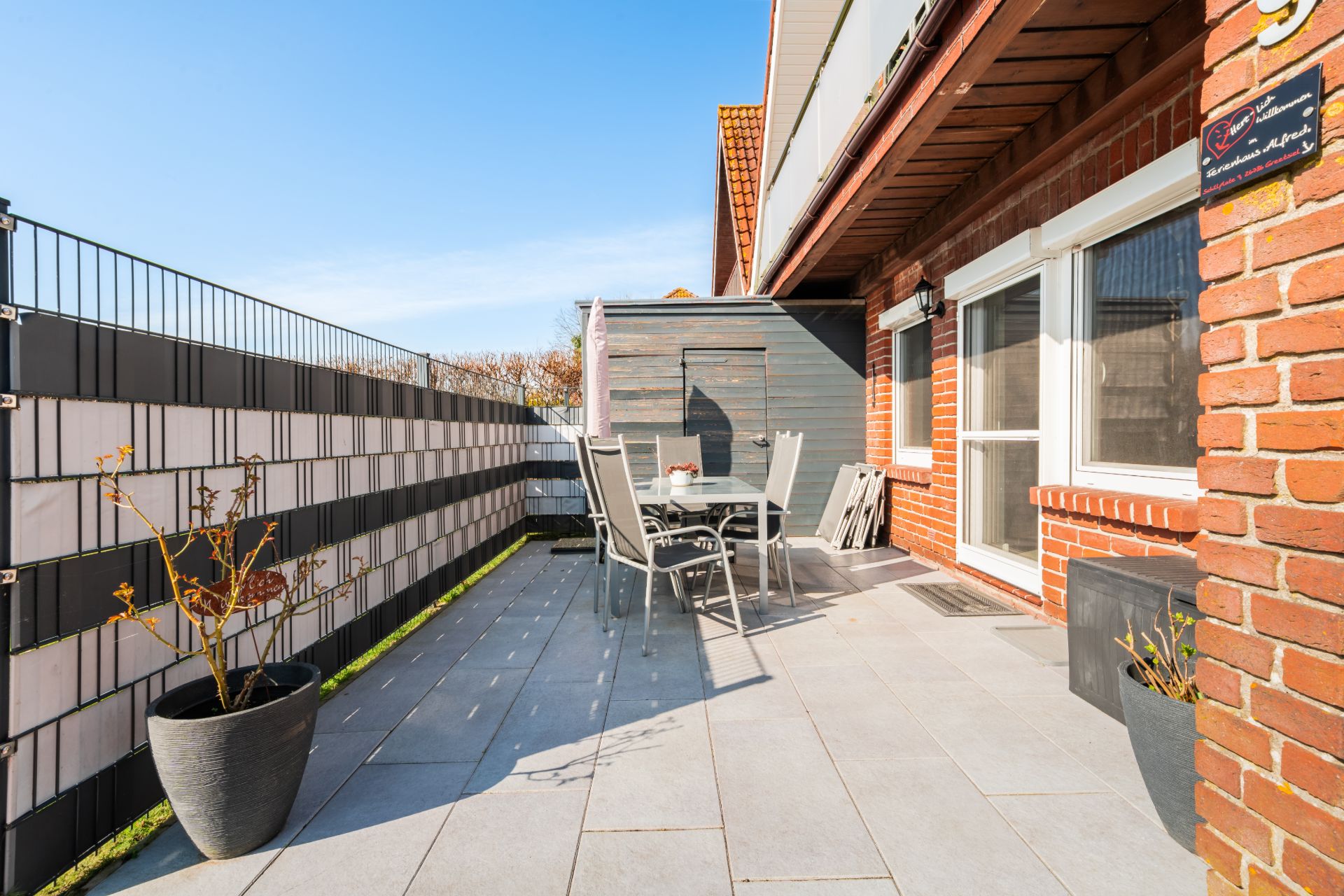 Große Terrasse mit Tisch, Stühlen, zwei Pflanzen und Sichtschutzzaun an einem Wohnhaus.