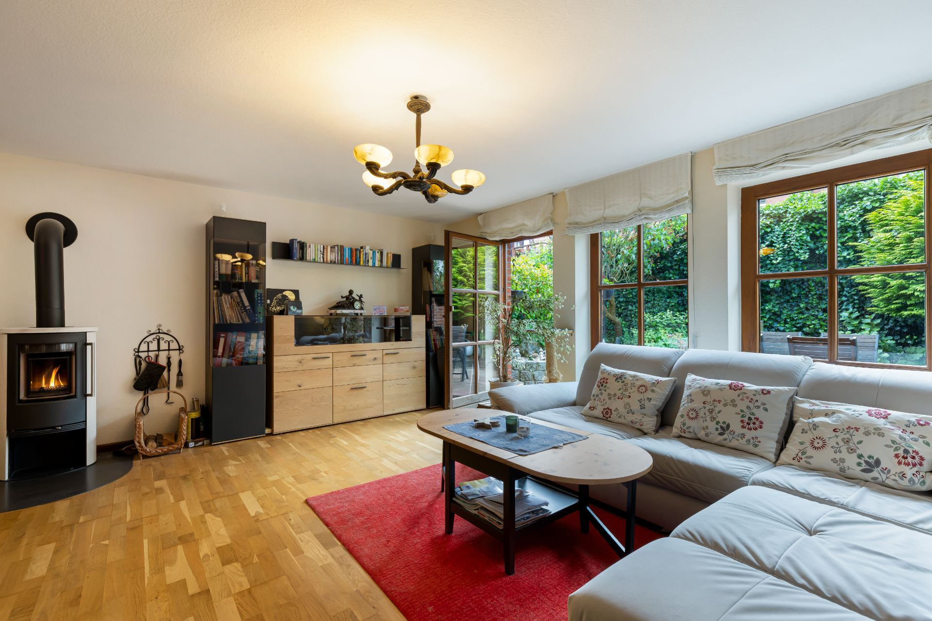 Wohnzimmer mit Ecksofa, Holzofen, Bücherregalen und großen Fenstern mit Blick in den Garten.