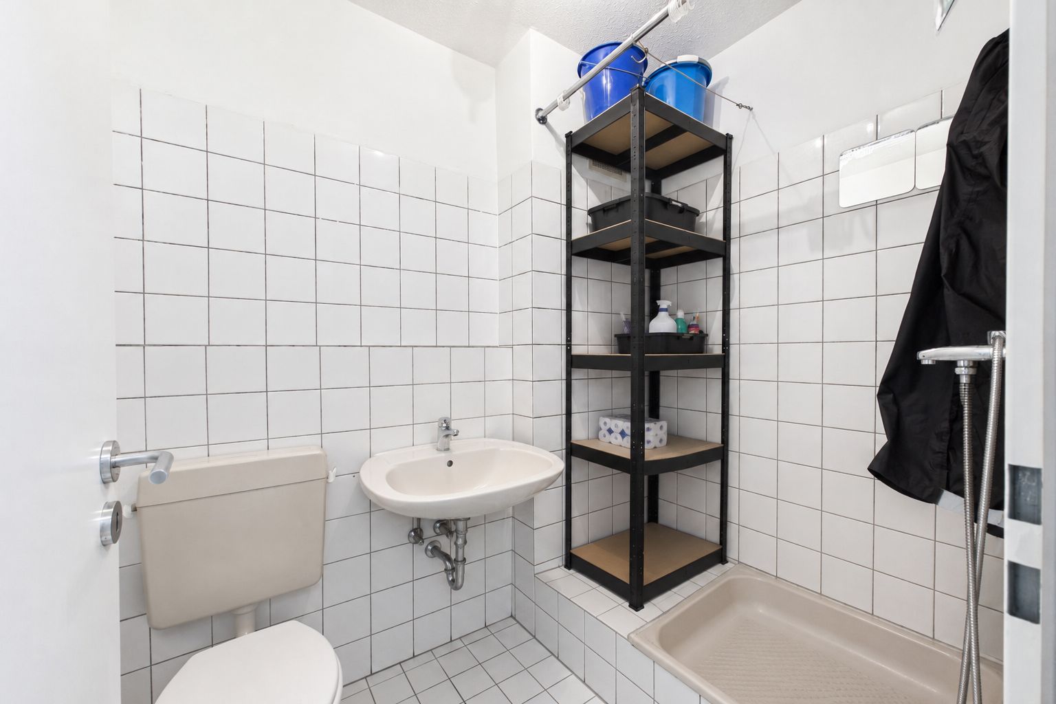 Kleines Badezimmer mit Toilette, Waschbecken, Duschwanne und Regal mit Haushaltsartikeln.