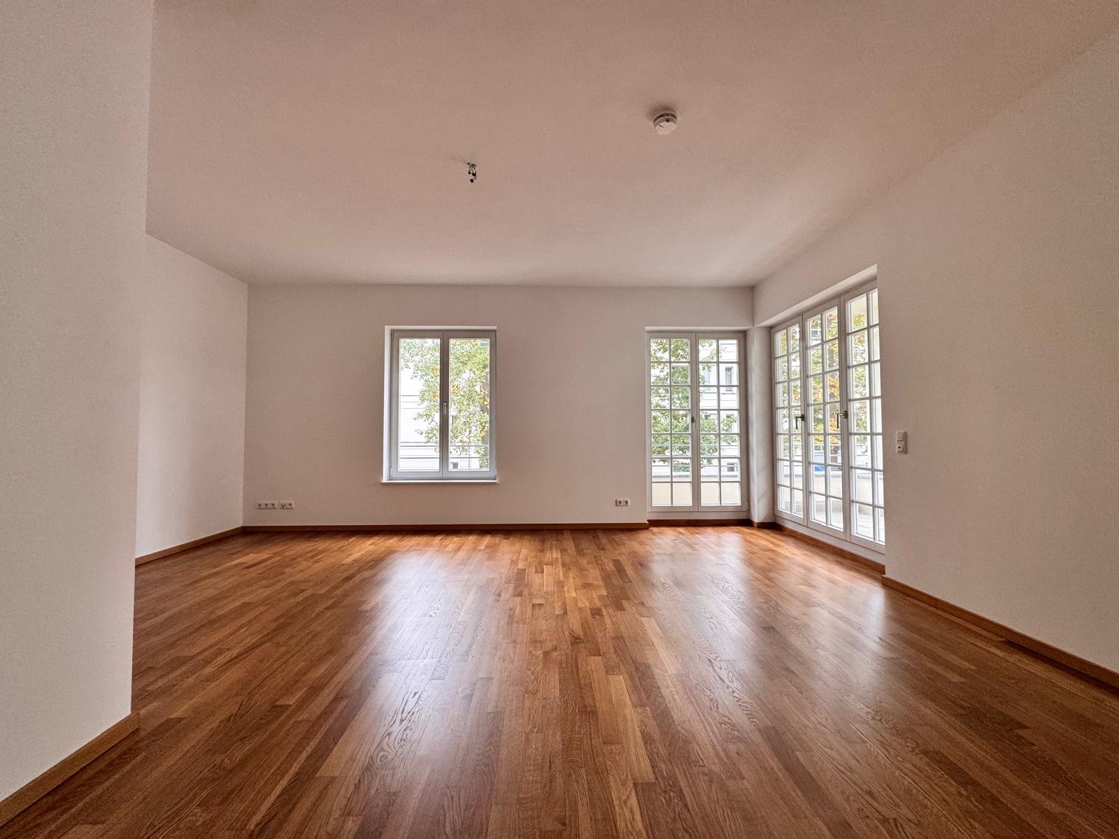 Helles, leeres Zimmer mit Parkettboden und großen Fenstern.