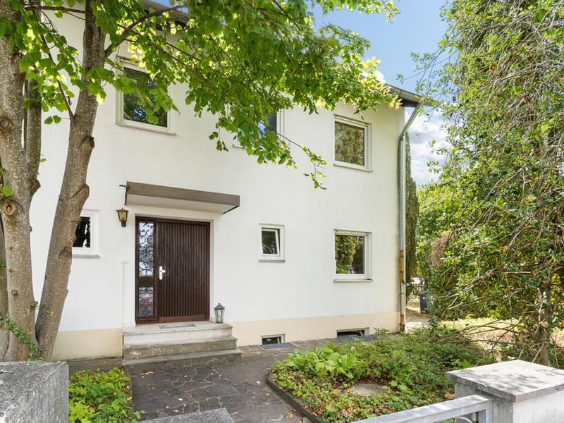 Eingangsbereich eines weißen Wohnhauses mit Garten und Baum im Vordergrund.