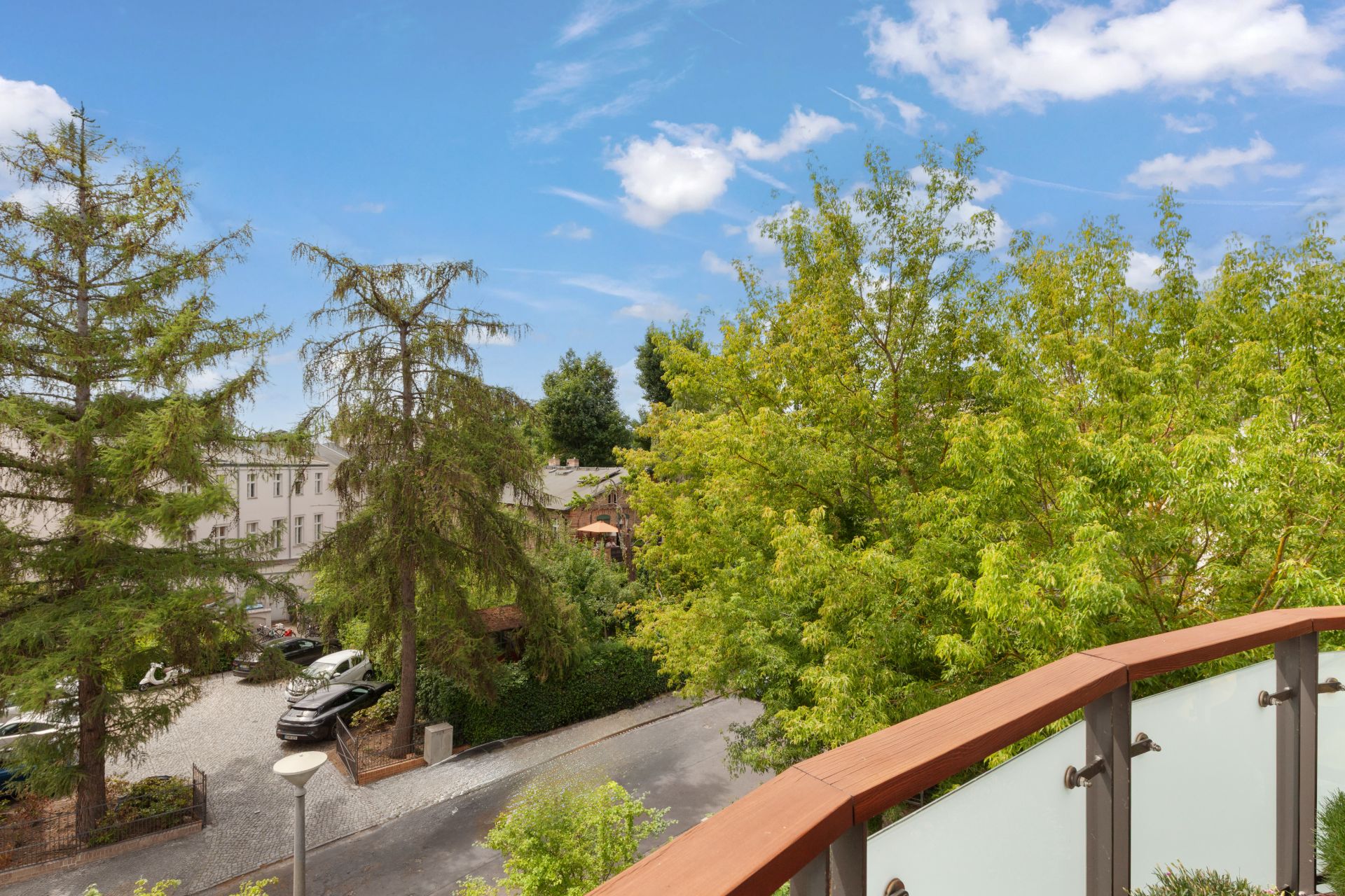 Blick vom Balkon auf eine grüne Baumlandschaft und eine ruhige Straße mit geparkten Autos.