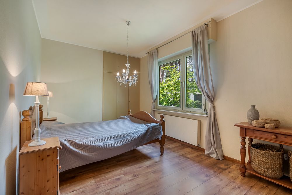 Helles Schlafzimmer mit Holzbett, Nachttisch, Fenster mit Vorhängen und Holztisch mit Körben.